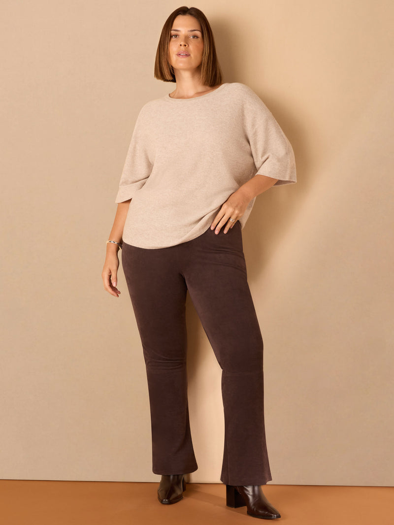 Woman wearing a beige sweater and brown pants against a beige wall.
