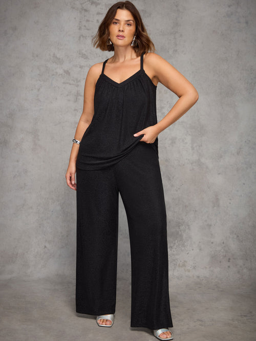 Woman wearing a black cami and sparkle trousers against a gray background