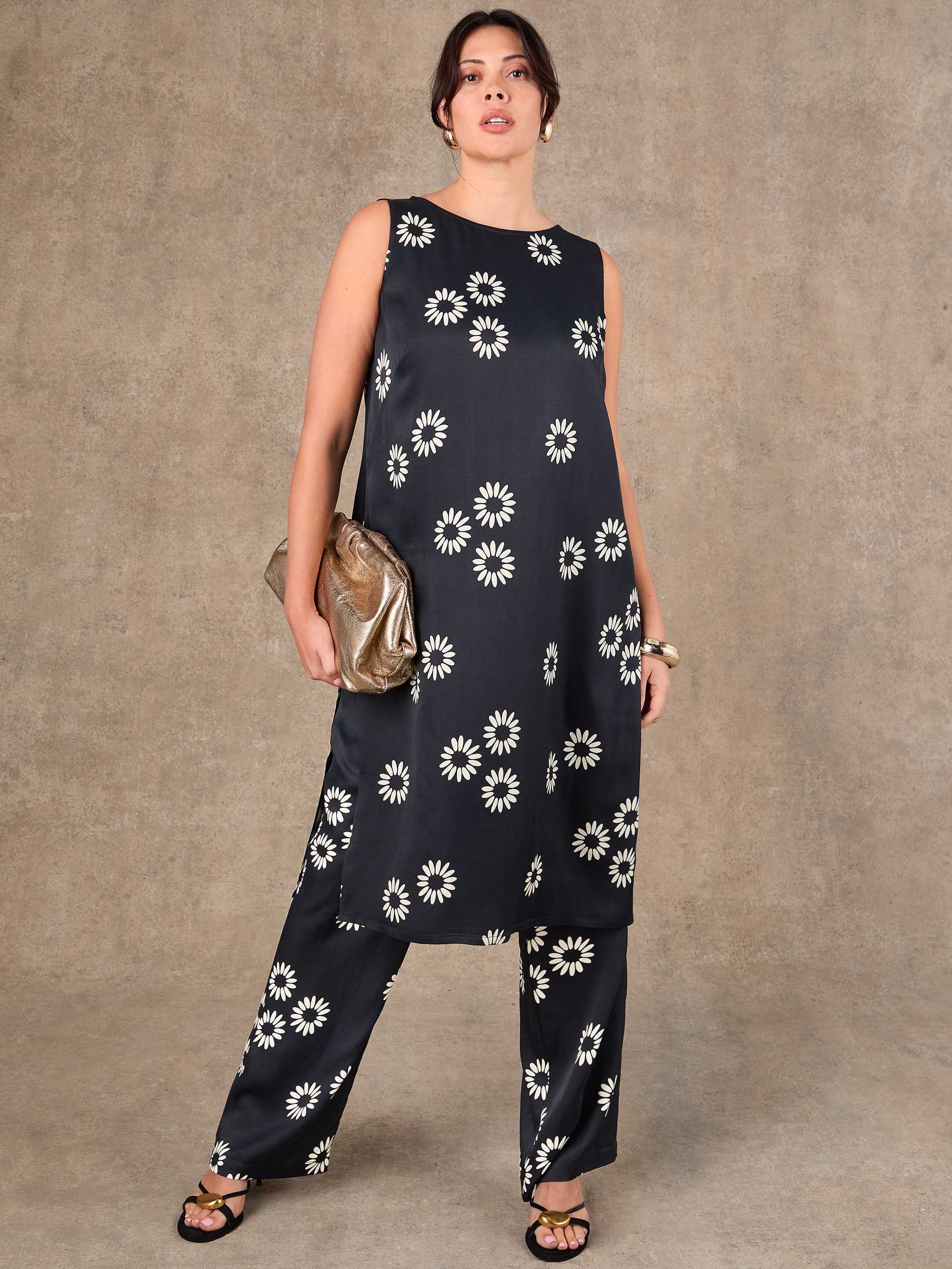 Woman wearing a black sleeveless top and wide-leg pants with white floral patterns against a brown background.