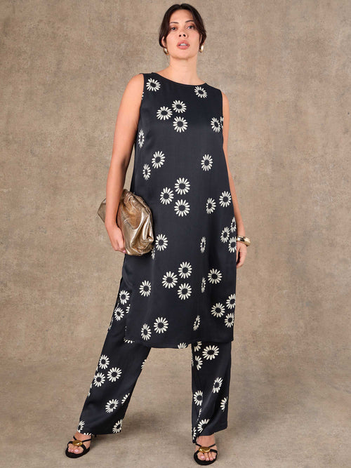 Woman wearing a black sleeveless top and wide-leg pants with white floral patterns against a brown background.